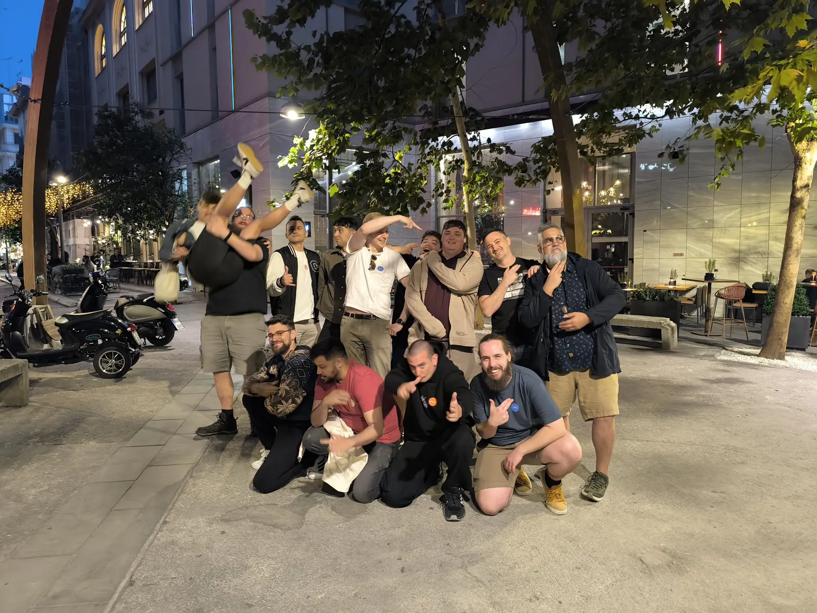 A corporate team strikes playful poses on a Thessaloniki street in the evening after a Salty Soil city treasure hunt, one teammate lifted upside down