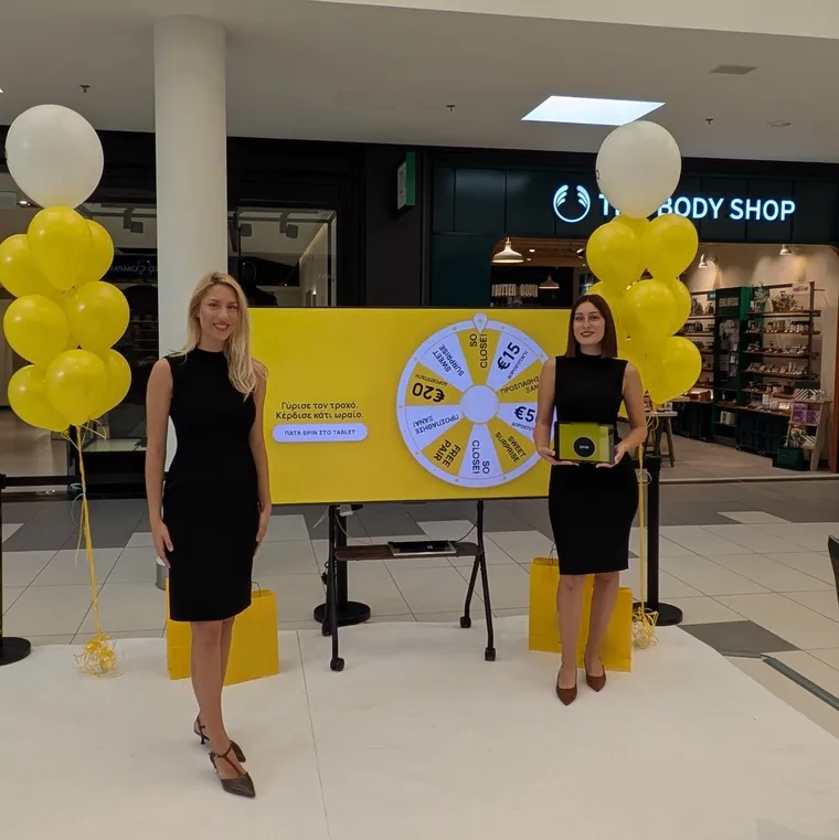 A branded prize-wheel activation with a 75-inch screen, an iPad spin button and two hosts at a store opening in a Thessaloniki shopping centre