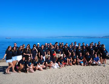 A large corporate group in matching T-shirts on a beach on the Thermaic Gulf, with Thessaloniki across the water, during a company retreat run by Salty Soil