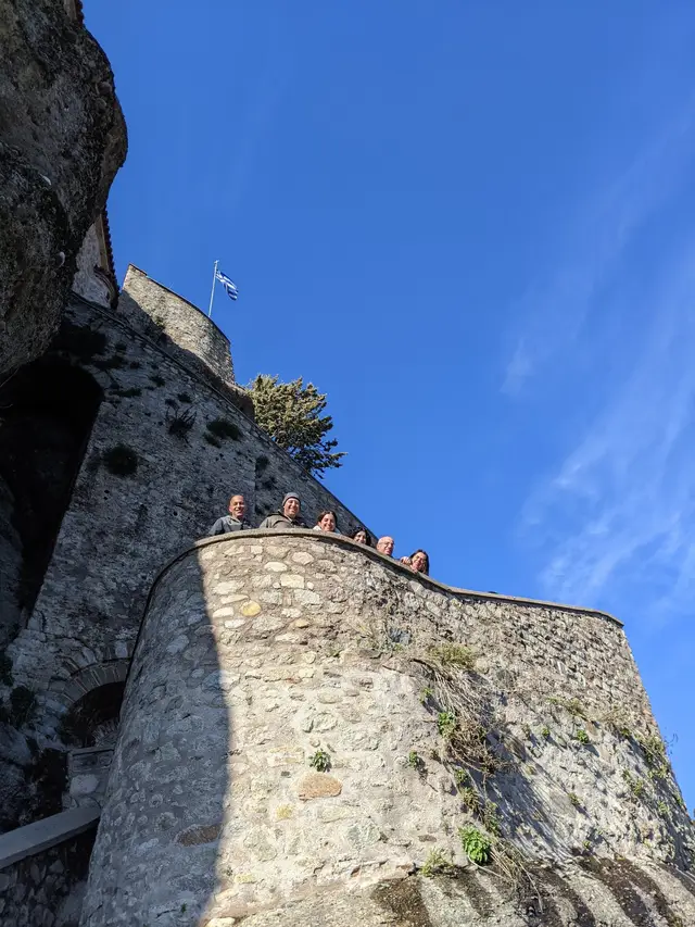 Guests look down from a monastery wall with a Greek flag above them at Meteora, on a Salty Soil day trip from Thessaloniki