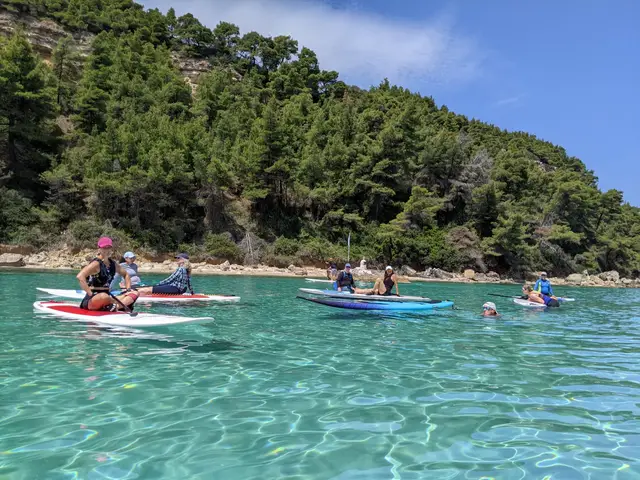 A group rests on their SUP boards in a turquoise cove below pine forest in Halkidiki, on a Salty Soil SUP trip