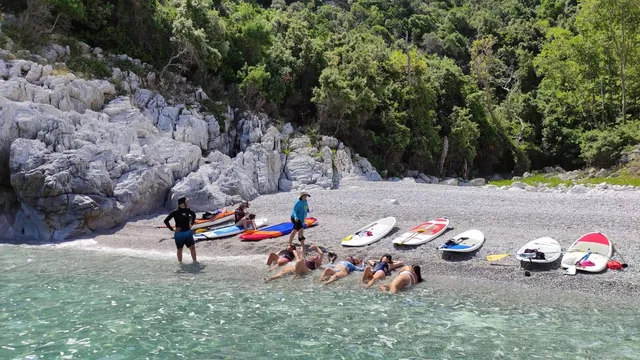 Κωπηλάτες χαλαρώνουν στα ρηχά μιας παραλίας με βότσαλα κάτω από λευκά βράχια και δάσος, με τις σανίδες SUP παραταγμένες στην ακτή, σε πολυήμερη εκδρομή SUP της Salty Soil