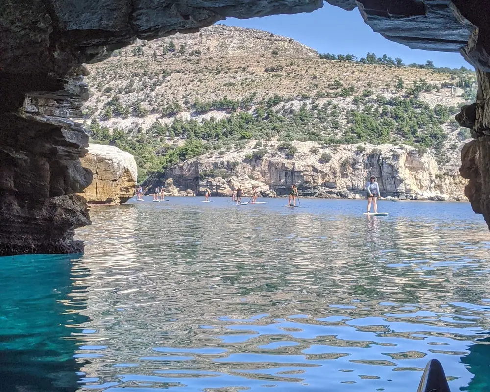 Ομάδα SUP ιδωμένη μέσα από την αψίδα θαλάσσιας σπηλιάς, σε κρυστάλλινα τιρκουάζ νερά, σε πολυήμερη εκδρομή SUP της Salty Soil στην Ελλάδα