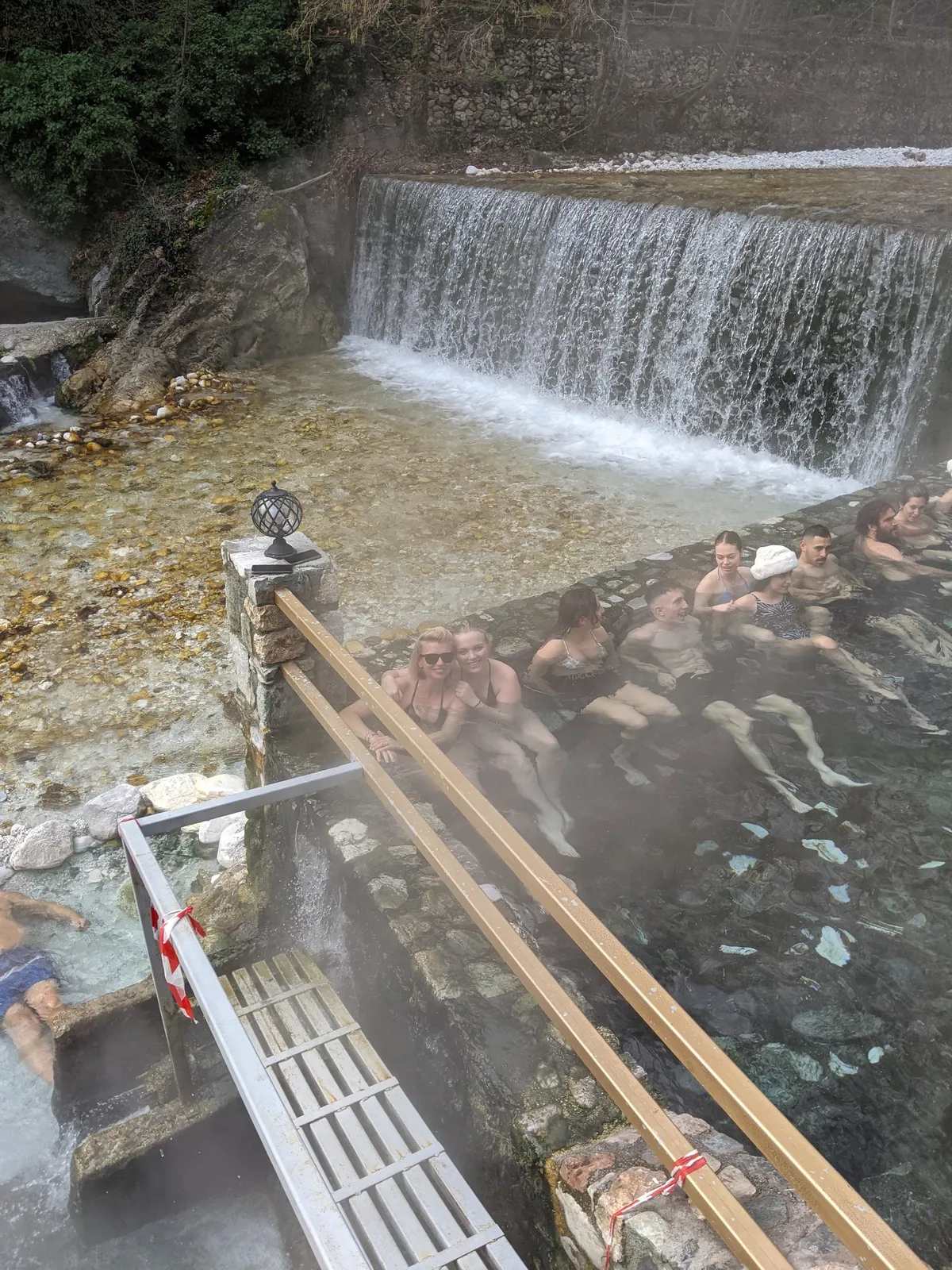 Λουόμενοι χαλαρώνουν στην αχνιστή υπαίθρια ιαματική πισίνα κάτω από έναν καταρράκτη στα Λουτρά Πόζαρ, σε ημερήσια εκδρομή της Salty Soil από τη Θεσσαλονίκη