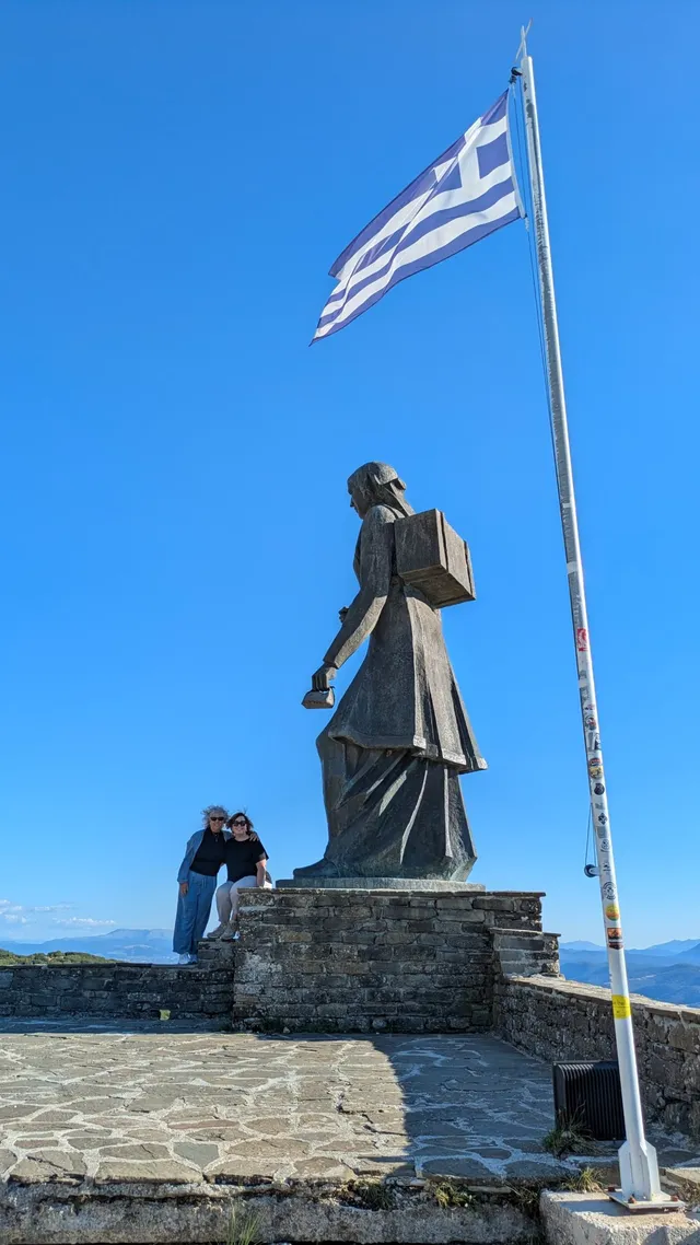 Two guests beside the bronze Monument to the Women of Pindus and a Greek flag in Epirus, on a Salty Soil private multi-day tour