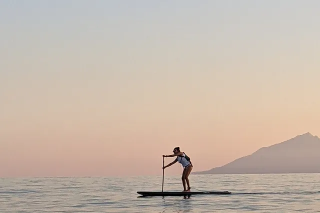 Κωπηλάτης σε σανίδα SUP σε ήρεμη θάλασσα στο ηλιοβασίλεμα, με το Άγιον Όρος στον ορίζοντα, σε ημερήσια εκδρομή SUP της Salty Soil στη Χαλκιδική