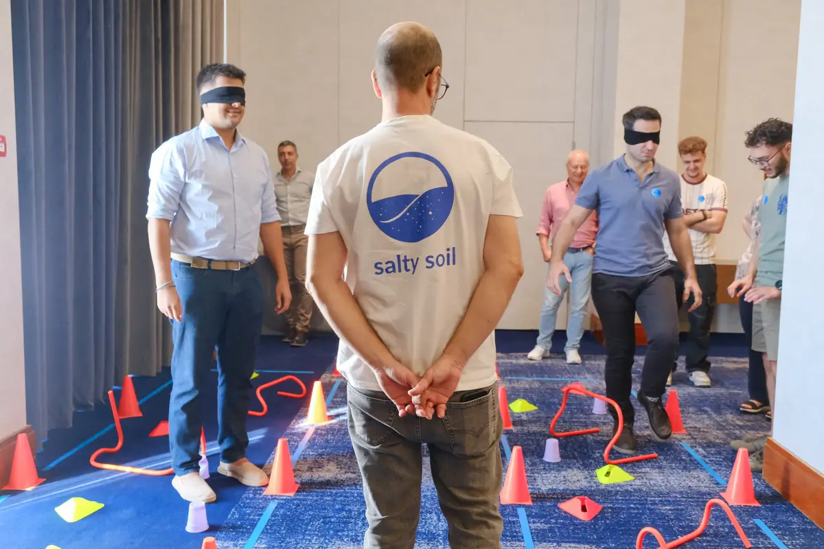 Blindfolded colleagues cross an obstacle course of cones and hurdles while a Salty Soil facilitator watches, during an indoor team building session in Thessaloniki