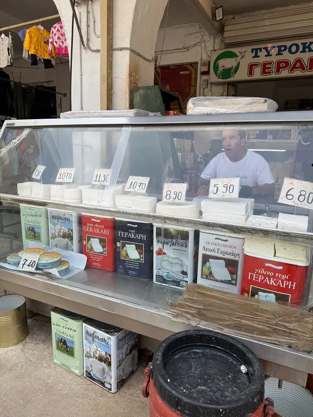 A cheesemonger behind a counter of Greek white cheeses and feta tins at a market shop in Thessaloniki, a stop on the Salty Soil food tour