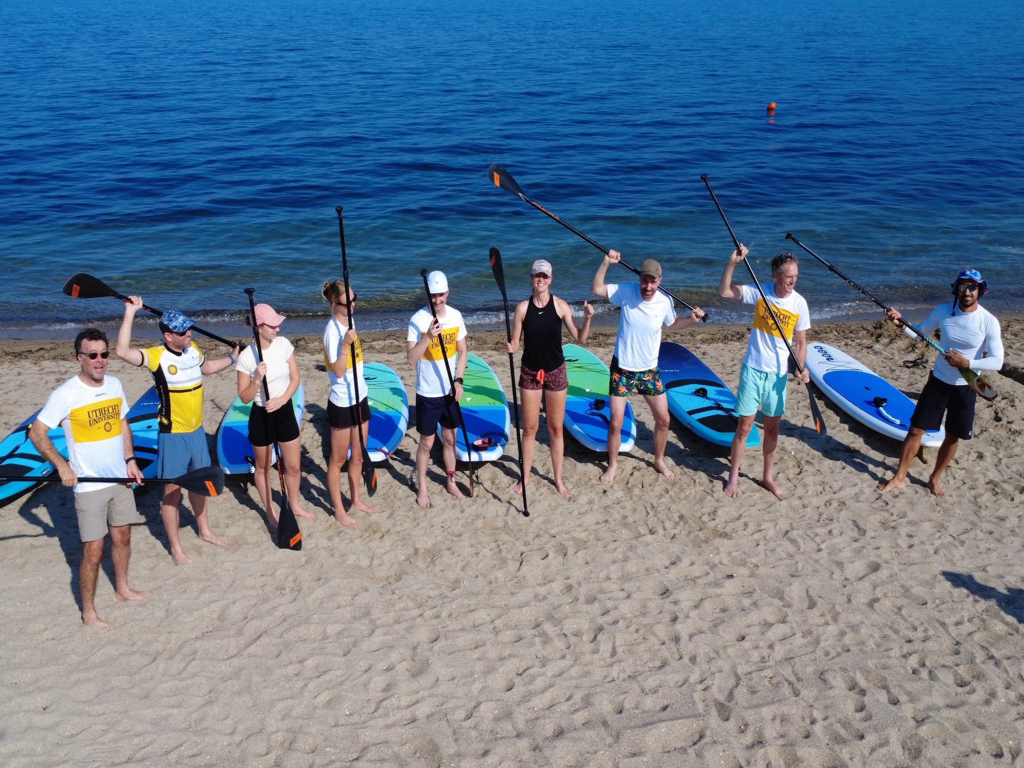 Group of nine paddlers holding paddles beside SUP boards on the sandy beach at Perea, Thessaloniki, before a Salty Soil stand-up paddleboard tour